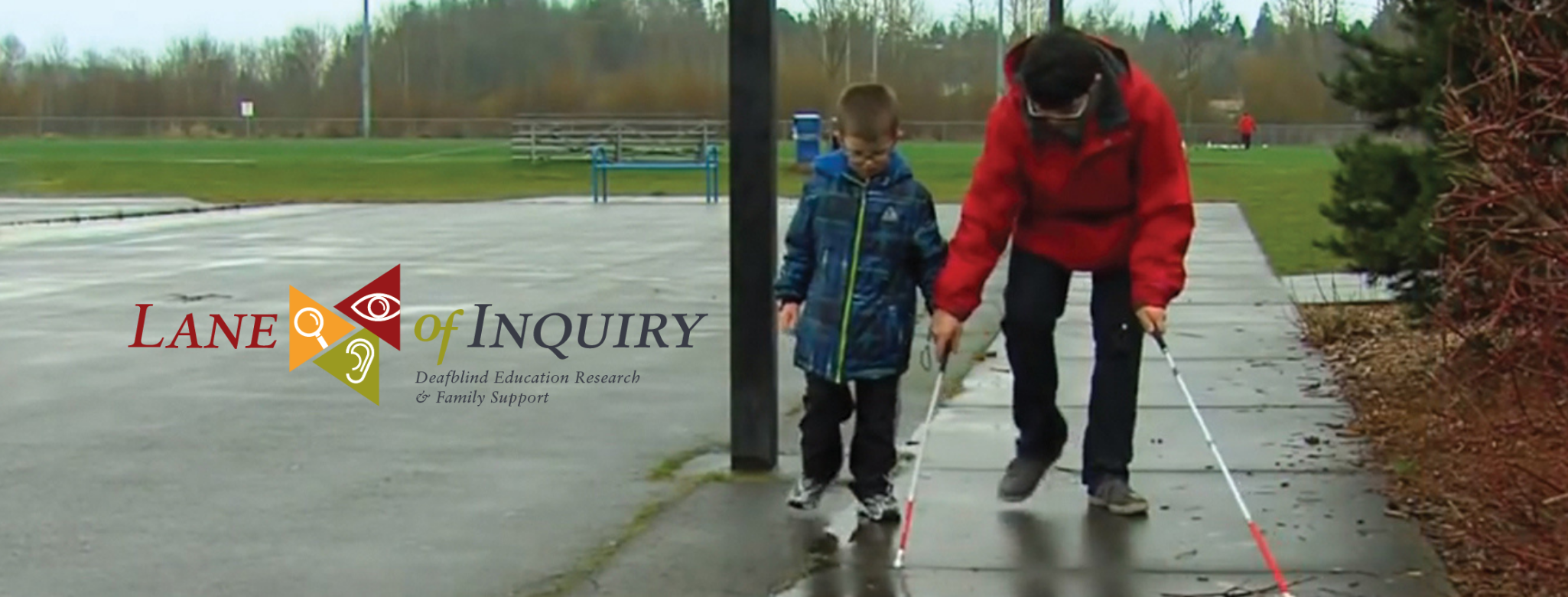 Older boy on right in red rain coat helping younger boy on left in blue rain coat learn how to use a white cane.
