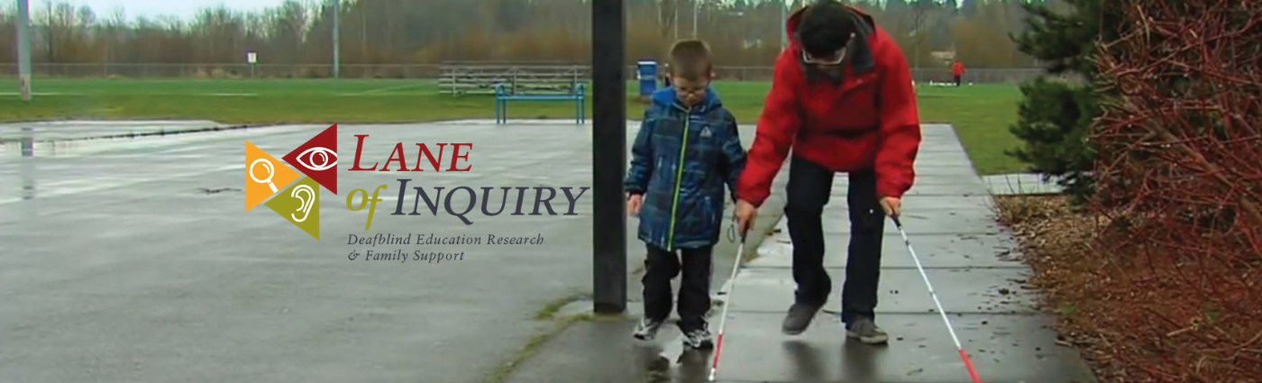 Older boy on right in red rain coat helping younger boy on left in blue rain coat learn how to use a white cane.