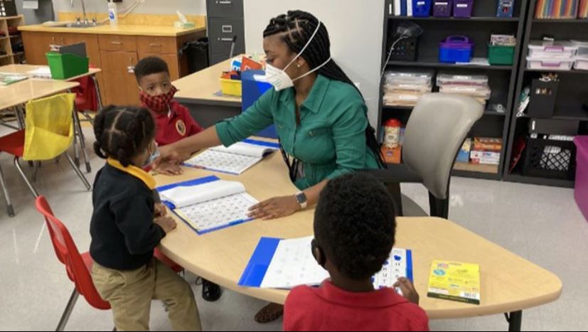 Black teacher seated at desk with 3 young Black students with open workbooks